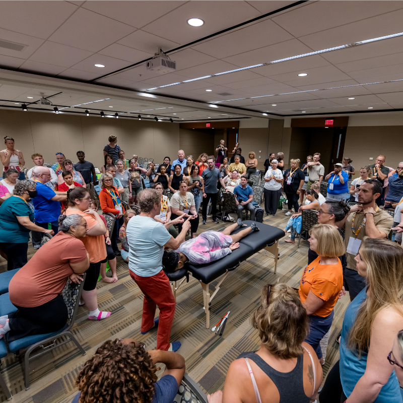 An image of a massage therapist demonstrating a massage technique while a client lays on a massage table; a large crowd of viewers surround him.