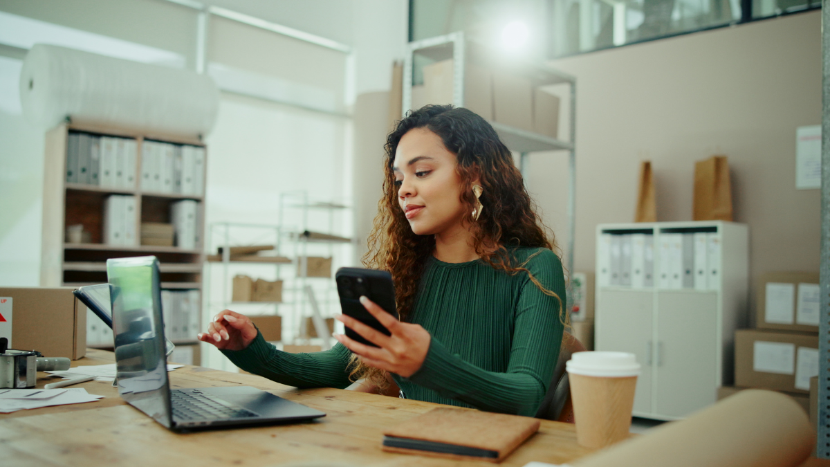 An image of a woman with curly hair sitting at a desk comparing information on her phone and a computer. She is in an office setting with a notebook by her side.