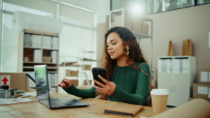 Image of a woman with curly hair sitting at a desk comparing information on her phone and a laptop, an office setting is in the background and a notebook next to her on the desk.