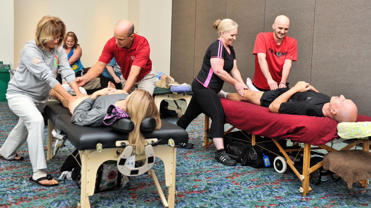 An image of a group of massage therapists: there are two tables with clients on them, and two massage therapists are working with each client. Everyone is smiling as the therapists learn a new technique.
