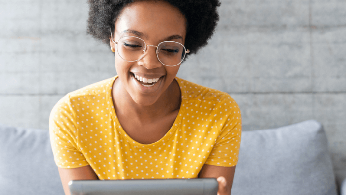 Image of a woman with glasses and a yellow shirt smiling and looking at something on a tablet.