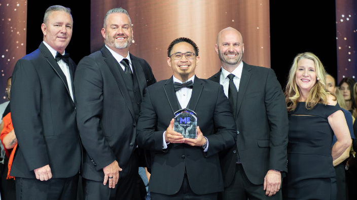An image of five people in black-tie attire, smiling. A man in the middle holds a trophy.