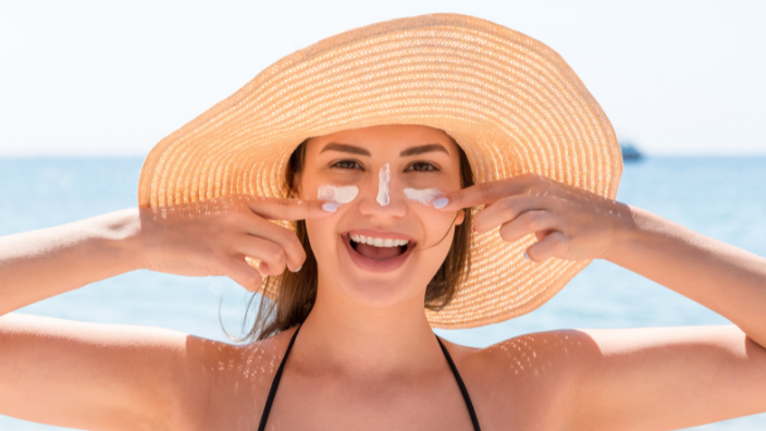 An image of a woman smiling in a wide-brimmed hat and sunscreen on her face at the beach.