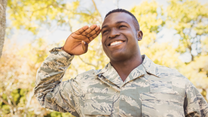 An image of a man in a military uniform saluting, trees are bloomed in the background.