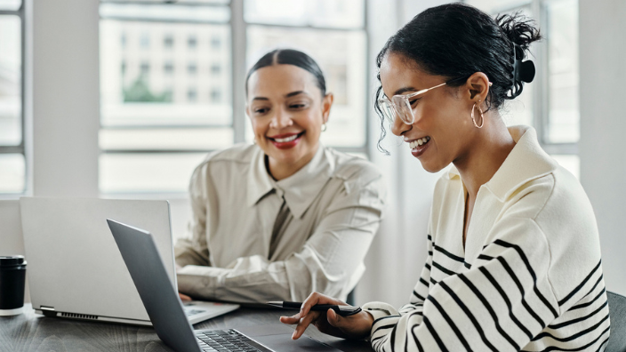 An image of two women sitting in an office, smiling with their laptops in front of them.